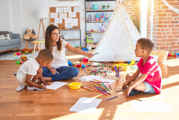 Jovem e bela professora e crianças sentadas no chão desenhando em torno de muitos brinquedos no jardim de infância
 - Foto, Imagem