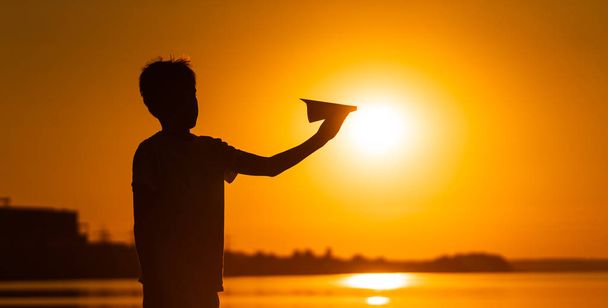 Black silhouette of a boy standing against orange summer sunset background. holding small paper plane in hands. Horizontal photo. - Fotoğraf, Görsel