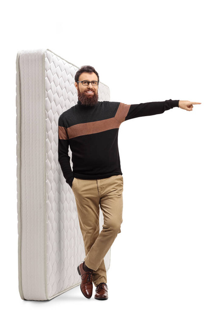Bearded man leaning on a bed mattress and pointing to the side - 写真・画像