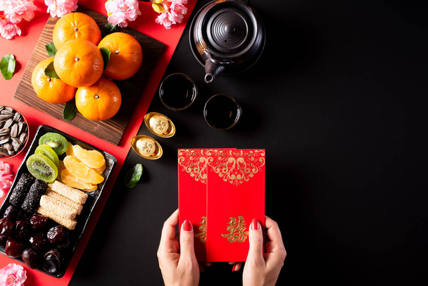 Chinese new year festival decorations. Woman hand holding pow or red packet, orange and gold ingots on a black stone background. Chinese characters FU means fortune good luck, wealth, money flow. - Foto, afbeelding