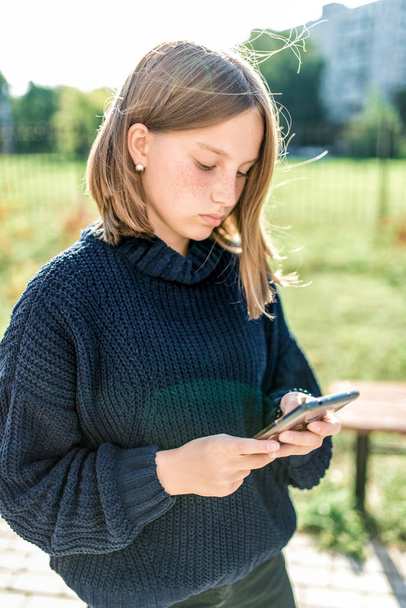 teenager girl 14-15 years old, autumn day, stands on street city, holds smartphone her hands, reads and writes message an application, online to Internet. Social networks. Casual warm clothes sweater. - Foto, imagen