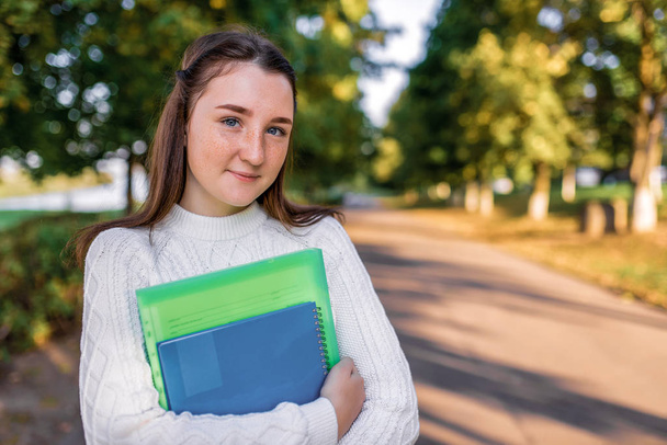 Happy teenager girl smiling, standing in summer park, autumn trees background, free space copy text. In hands notebook textbooks and notes, preparation for school college. Warm clothes sweater. - Photo, Image