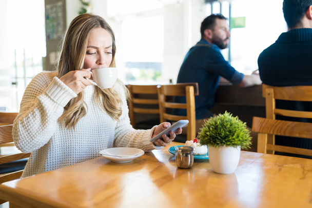 Νεαρή γυναίκα πίνοντας καφέ, ενώ γραπτών μηνυμάτων στο smartphone στο τραπέζι στο καφέ - Φωτογραφία, εικόνα
