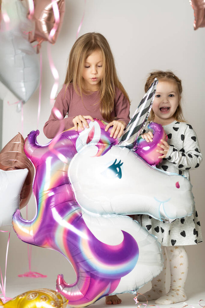Two little girls play with a big unicorn balloon surrounded by balloons - Фото, изображение