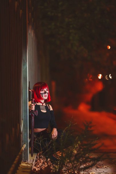 Ragazza cosplay a immagine di un elfo scuro in una parrucca rossa con una spada sullo sfondo della città di notte. Una donna con i capelli rossi in costume da elfo con orecchie finte e trucco. Abito elfo scuro per Halloween
. - Foto, immagini