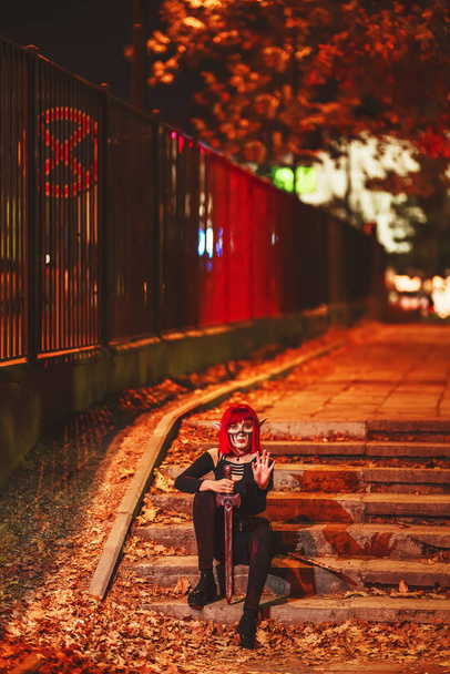 Cosplay menina na imagem de um duende escuro em uma peruca vermelha com uma espada no fundo da cidade noturna. Uma mulher com cabelo ruivo num traje de duende com orelhas falsas e maquilhagem. Roupa de elfo escuro para Halloween
. - Foto, Imagem