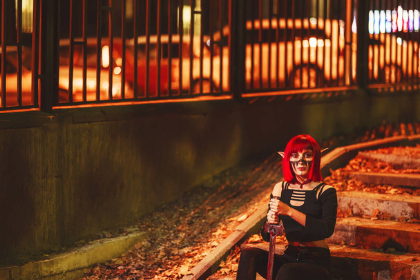 Девушка-косплей в образе темного эльфа в красном парике с мечом на фоне ночного города. Женщина с рыжими волосами в костюме эльфа с фальшивыми ушами и макияжем. Тёмный костюм эльфа на Хэллоуин
. - Фото, изображение