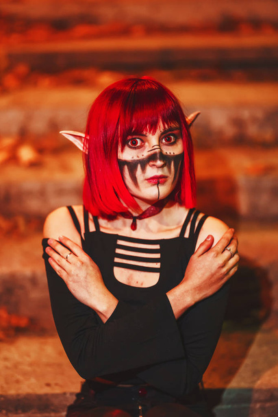 Cosplay menina na imagem de um duende escuro em um retrato de peruca vermelha close-up. Uma mulher com lentes vermelhas nos olhos e cabelos vermelhos em um traje de elfo com orelhas falsas e maquiagem. Roupa de elfo escuro para Halloween
. - Foto, Imagem