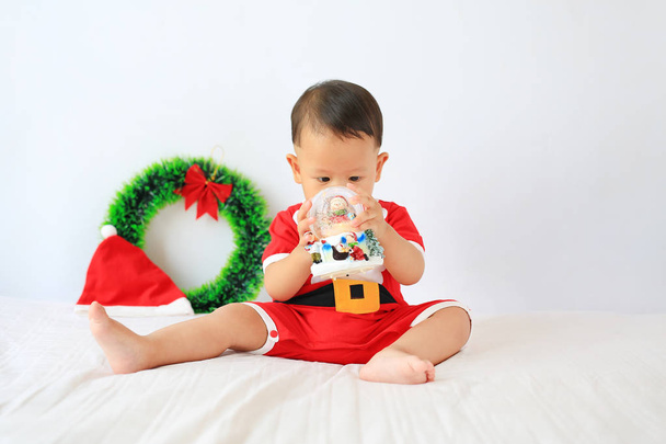 Little Asian baby boy in Santa costume with holding snow globe sitting on bed over white background. Merry Christmas and Happy New Year Concept. - Foto, afbeelding