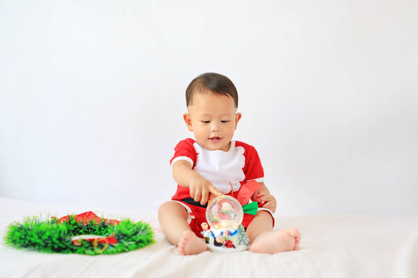 Little Asian baby boy in Santa costume playing snow globe sitting on bed over white background. Merry Christmas and Happy New Year Concept. - Φωτογραφία, εικόνα