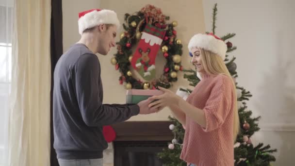 Boční pohled na mladou bělošku v červeném vánočním klobouku, jak se dívá dovnitř dárku, kterou jí dal manžel a dělá nespokojený obličej. Nešťastná dívka zklamaná dárkem. Sváteční sezóna. - Záběry, video