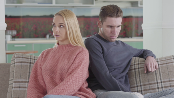Portrait of upset Caucasian couple sitting on couch back to back and thinking. Sad young woman and man argued. Relationship, communication, problems. - Felvétel, videó