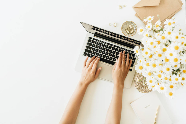 Women's hands work composition. Home office desk workspace with laptop, chamomile daisy flowers bouquet and notebook on white background. Flat lay, top view freelancer concept with copy space mockup. - Photo, Image
