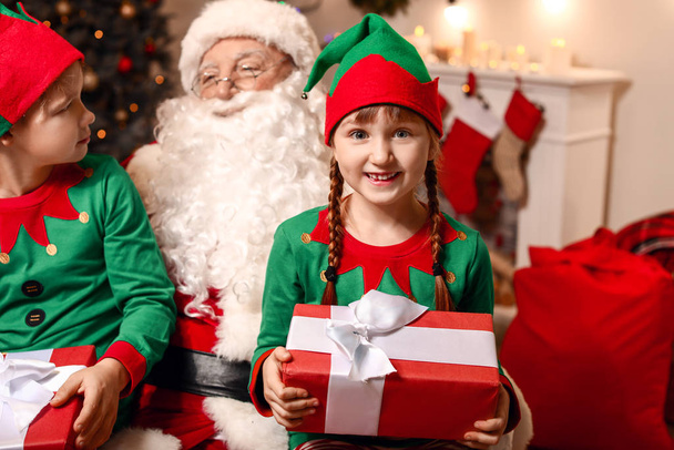Papai Noel e pequenas crianças elfo com presentes no quarto decorado para o Natal
 - Foto, Imagem