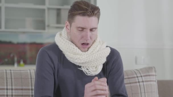 Close-up of Caucasian man in white scarf sitting on sofa and sneezing. Portrait of sick guy at home. Illness, healthcare and medicine, family care. - Felvétel, videó