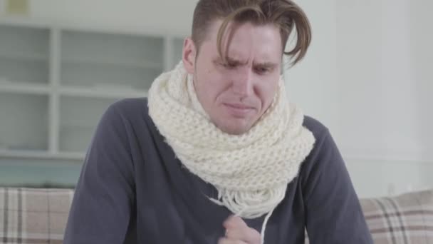 Close-up portrait of Caucasian man in white scarf sitting on sofa, sneezing and coughing. Portrait of sick guy shivering at home. Illness, healthcare and medicine. - Footage, Video