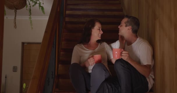 Front view of a Caucasian couple at home, sitting side by side on the stairs holding cups of coffee, talking and kissing, slow motion 4k - Πλάνα, βίντεο