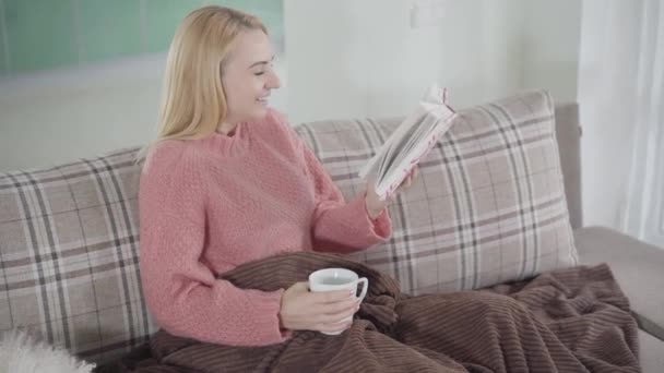 Cámara acercándose a la risa mujer caucásica leyendo libro en casa. Hermosa mujer sentada con novela y taza de té caliente o café en el sofá. Aficion, ocio, estilo de vida
. - Imágenes, Vídeo