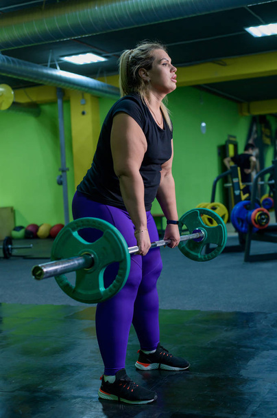 Fat European woman is engaged in fitness with a barbell in the gym. A fat girl does deadlift. - Foto, afbeelding