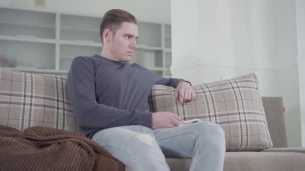Portrait of angry Caucasian man sitting on couch with smartphone. Young guy waiting nervously for wife or girlfriend at home. Relationship problems. - Séquence, vidéo