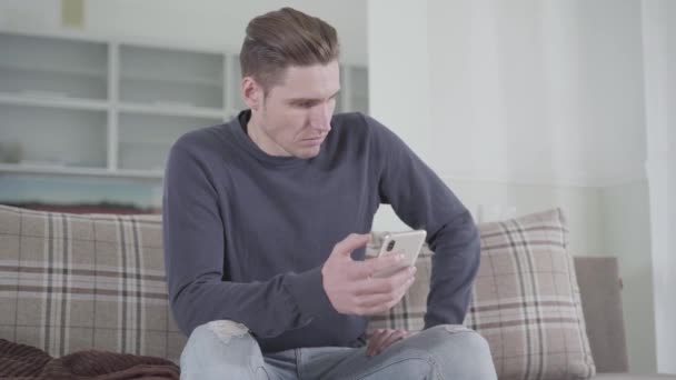 Portrait of angry Caucasian man looking at smartphone screen and beating fist against knee. Young guy waiting nervously for wife or girlfriend at home. Relationship problems. - Séquence, vidéo