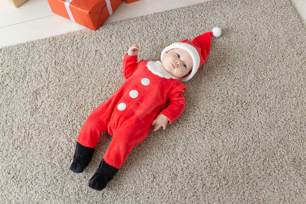 Piękne maleństwo świętuje święta. Noworoczne wakacje. Śmieszne dziecko w stroju świątecznym i w czapce Świętego Mikołaja, widok z góry - Zdjęcie, obraz