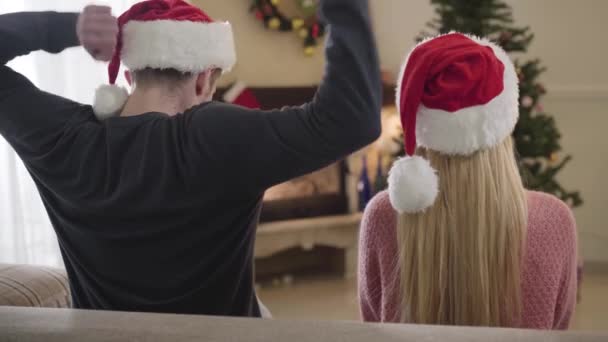 Back view of adult Caucasian man stretching and putting hand on womans shoulder. Couple in New Year hats sitting on couch in front of Christmas tree. Holidays season, love, relationship. - Filmagem, Vídeo