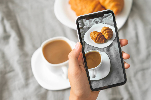 Mano femenina con teléfono móvil tomando fotos de sabroso desayuno café y croissants
  - Foto, imagen