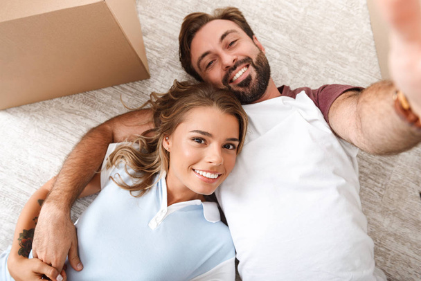 Photo of happy couple taking selfie photo and lying near cardboard box - Foto, Imagem