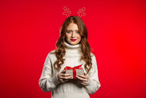 Young pretty woman smiling and holding gift box on red studio background. Girl with beautiful hairstyle in white sweater and deer horns. Christmas mood. - Фото, зображення