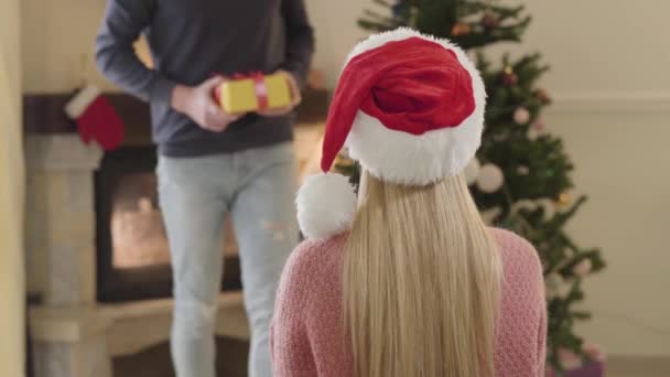 Young Caucasian man coming from the background with wrapped gift, excited woman shaking present and hugging guy. Happy smiling family spending holidays together at home. - Filmati, video