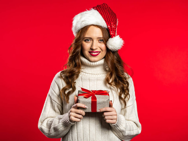 Young pretty woman smiling and holding gift box on red studio background. Girl with beautiful hairstyle in white sweater and Santa hat. Christmas mood. - Фото, изображение