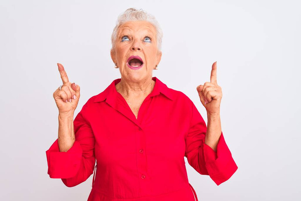 Senior mulher de cabelos grisalhos vestindo camisa casual vermelho em pé sobre fundo branco isolado espantado e surpreso olhando para cima e apontando com os dedos e braços levantados
. - Foto, Imagem