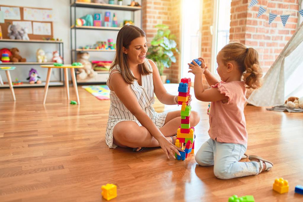 Красивая учительница и блондинка девочка строит башню, используя пластиковые блоки в детском саду
 - Фото, изображение