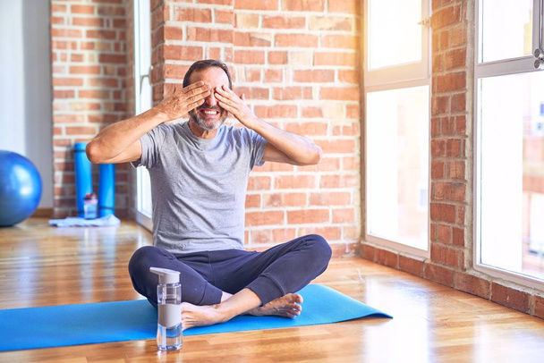Przystojny sportowiec w średnim wieku siedzi na macie i ćwiczy jogę na siłowni, przykrywając oczy dłońmi uśmiechniętymi wesołymi i zabawnymi. Koncepcja niewidomych. - Zdjęcie, obraz