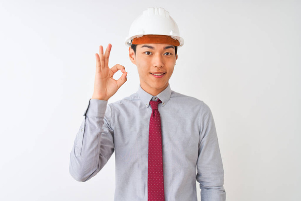 Китайский архитектор мужчина в галстуке и шлеме стоит на изолированном белом фоне улыбаясь позитивно делает хорошо знак с рукой и пальцами. Успешное выражение
. - Фото, изображение