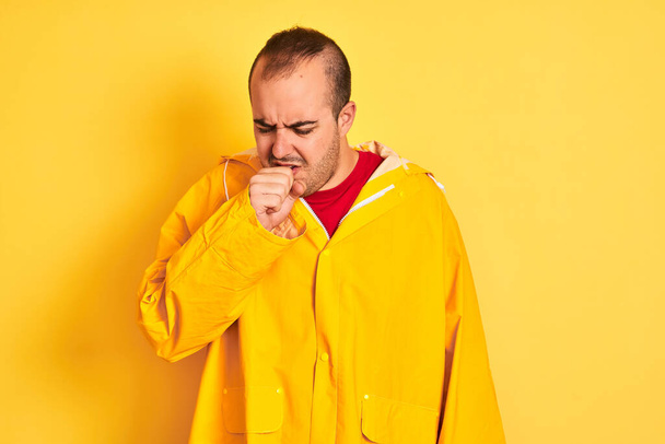 junger Mann im Regenmantel, der vor isoliertem gelben Hintergrund steht, fühlt sich unwohl und hustet als Symptom für Erkältung oder Bronchitis. Gesundheitskonzept. - Foto, Bild