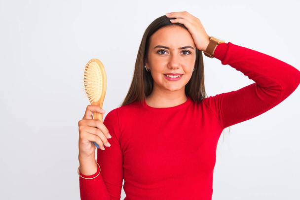 Young beautiful girl holding hair comb standing over isolated white background stressed with hand on head, shocked with shame and surprise face, angry and frustrated. Fear and upset for mistake. - Foto, Imagem