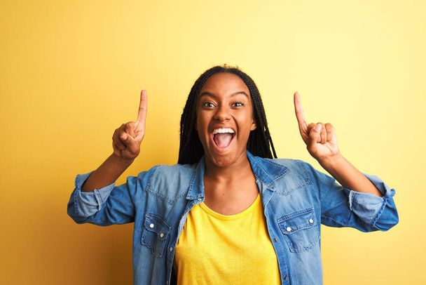 Νεαρή Αφροαμερικανή γυναίκα που φοράει τζιν πουκάμισο στέκεται πάνω από απομονωμένο κίτρινο φόντο χαμογελώντας έκπληκτος και δείχνοντας προς τα πάνω με τα δάχτυλα και σήκωσε τα χέρια. - Φωτογραφία, εικόνα