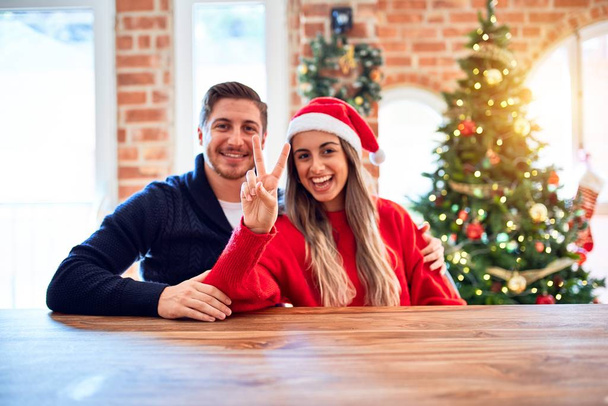 Jeune couple portant santa claus chapeau assis sur la chaise et la table autour de l'arbre de Noël à la maison montrant et pointant vers le haut avec les doigts numéro deux tout en souriant confiant et heureux
. - Photo, image