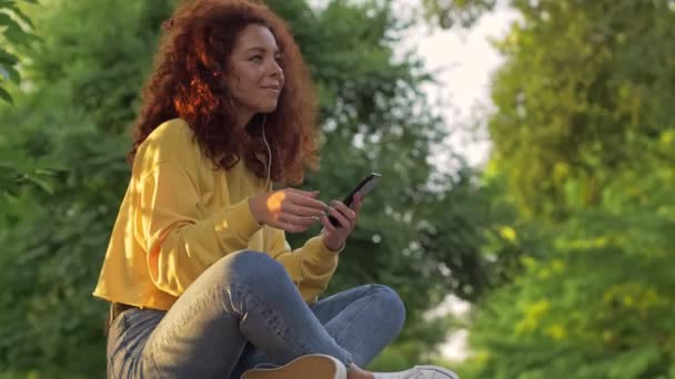 Cheerful young woman with curly redhead hair listening to music with smartphone and wired earphones while sitting on bench in green park - Filmmaterial, Video