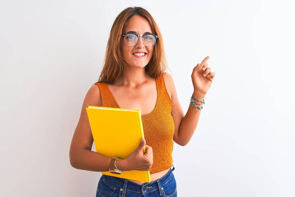 Young beautiful redhead woman wearing glasses reading a book over isolated background very happy pointing with hand and finger to the side - Photo, Image