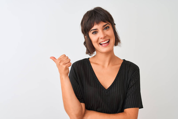 Young beautiful woman wearing black t-shirt standing over isolated white background smiling with happy face looking and pointing to the side with thumb up. - Photo, Image