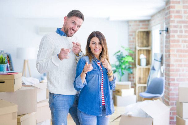 Jeune beau couple debout à la nouvelle maison autour des boîtes en carton pointant les doigts vers la caméra avec un visage heureux et drôle. Bonne énergie et vibes
. - Photo, image