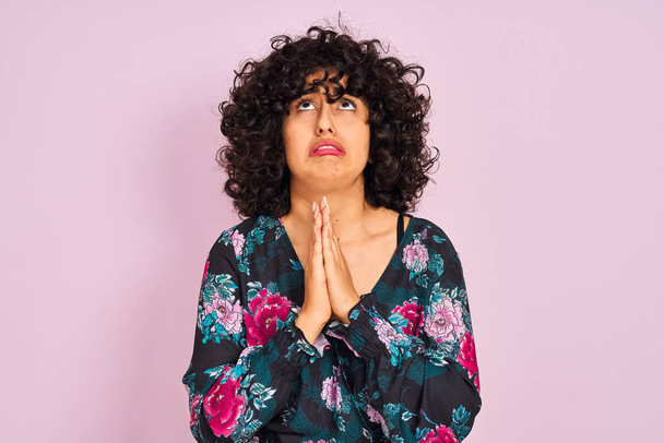 Mujer árabe joven con el pelo rizado con vestido floral sobre fondo rosa aislado mendigando y rezando con las manos junto con la expresión de esperanza en la cara muy emocional y preocupado. Pedir perdón. Concepto de religión
. - Foto, imagen