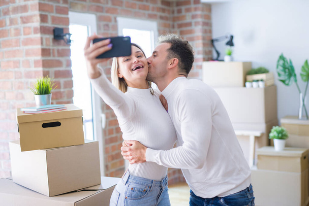 Молодая красивая пара стоит с помощью смартфона, чтобы сделать селфи поцелуи в новом доме вокруг картонных коробок
 - Фото, изображение