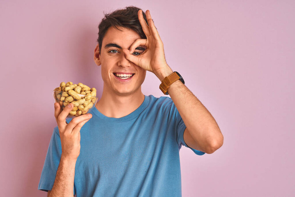 Adolescente menino segurando um monte de amendoins sobre fundo rosa isolado com o rosto feliz sorrindo fazendo ok sinal com a mão no olho olhando através dos dedos
 - Foto, Imagem