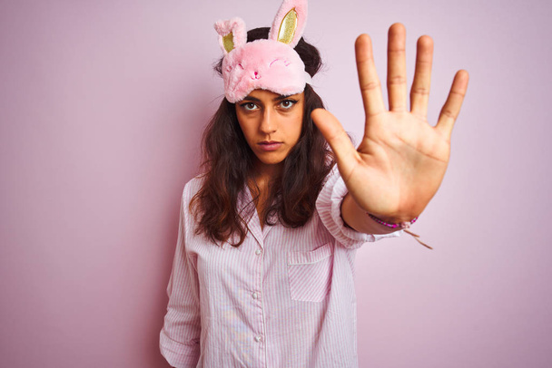 Young beautiful woman wearing pajama and sleep mask over isolated pink background doing stop sing with palm of the hand. Warning expression with negative and serious gesture on the face. - Photo, Image