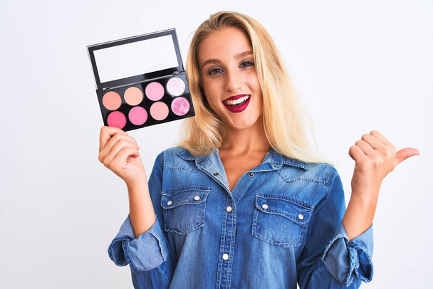 Young beautiful blond woman holding makeup standing over isolated white background pointing and showing with thumb up to the side with happy face smiling - 写真・画像