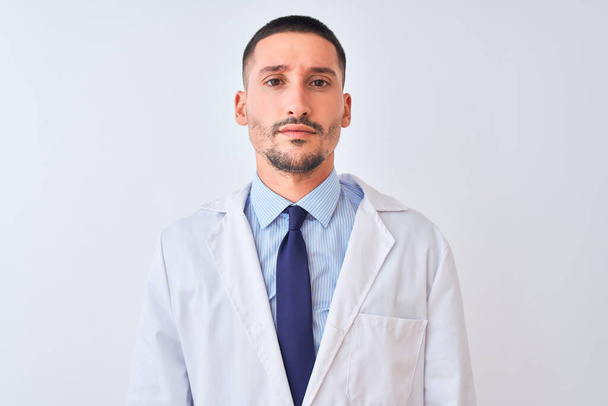 Homem jovem médico vestindo casaco branco sobre fundo isolado com expressão séria no rosto. Simples e natural olhando para a câmera
. - Foto, Imagem